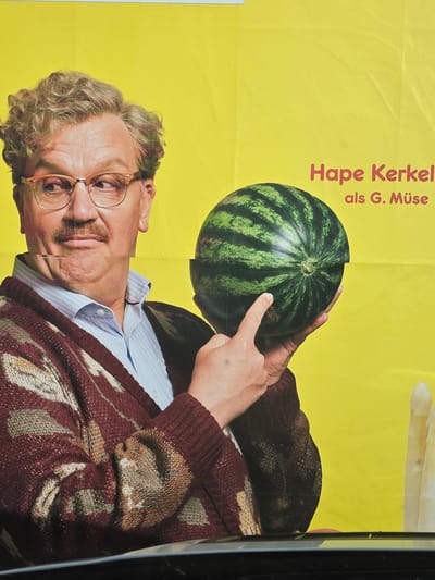 A large poster, located in Obergünzburg, Germany, displays a male subject against a solid bright yellow background. The man, depicted from approximately the chest up, possesses light brown curly hair, a prominent brown mustache, and wears orange-framed eyeglasses. He is dressed in a light blue and white striped button-up shirt underneath a dark red knitted cardigan featuring light-colored abstract patterns. His head is tilted slightly, and his eyes are directed towards his left, with a slightly open mouth suggesting a humorous or quizzical expression. He holds a whole, green-striped watermelon in his left hand, with his right index finger pointing precisely at a horizontal line traversing the watermelon's midsection, creating a visual segmentation. In the upper right quadrant of the poster, red text is visible, stating "Hape Kerkel" in a larger font above "als G. Müse" in a smaller font. To the right and slightly below the watermelon, a partial view of a white, elongated, and conical object, resembling white asparagus, is present. The poster material exhibits minor creasing and visible vertical seams. The lower edge of the poster is partially obscured by a dark, horizontal, reflective surface, possibly the top of a vehicle or a railing.