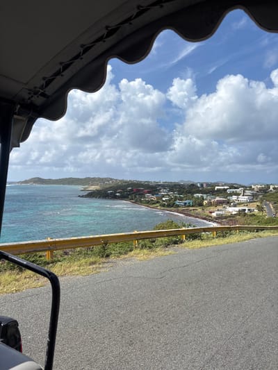 The image presents a panoramic view of Olde Yard Village, British Virgin Islands, captured from an elevated position within an open-air vehicle. In the upper foreground, a light-colored fabric canopy with a scalloped edge and visible lacing partially frames the sky. A black tubular metal support structure from the vehicle is visible on the lower left. The immediate midground features a paved asphalt road, exhibiting minor cracks, which descends gently towards the right. A yellow metal guardrail, showing some wear and discoloration, runs parallel to the road's edge, delineating the elevated pathway from the coastal drop-off. Green grass and low vegetation grow between the road and the guardrail, and along the hillside.
The dominant background comprises a tropical coastal landscape. The ocean displays clear turquoise waters near the shore transitioning to deeper blues further out, with visible waves breaking along the extensive reddish-brown beach. Numerous buildings, varying in size and color (including white, beige, blue, red, and green), are scattered along the coastline and extend up the verdant hillsides, forming the Olde Yard Village settlement. A distinct green headland juts into the sea on the left side of the bay. The sky is partially covered by large, white cumulus clouds against a clear blue background. No individuals or discernible activities are present in the scene.