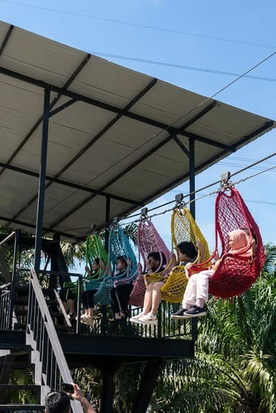 In Bintang Meriah, Indonesia, a group of five young individuals, four females and one male, are seated in colorful woven swings suspended from a high-tension wire. The swings are red, yellow, pink, blue, and green. The individuals are dressed casually, with some wearing headscarves. They are positioned on a raised platform with a corrugated metal roof supported by black metal beams. A metal staircase is visible on the left side of the platform. The background consists of lush green palm trees under a clear blue sky, suggesting an outdoor recreational setting. A person in the foreground is taking a photograph of the scene with a mobile phone.