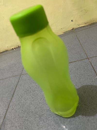 A lime green plastic water bottle with a matching green cap is positioned diagonally on a tiled floor. The floor tiles are gray and appear to be square. Behind the bottle, a yellow wall is visible, showing signs of wear and some dark discoloration along the bottom edge where it meets the floor. The scene is lit, casting a shadow of the bottle to the right. The location is described as Anggaswangi, Indonesia, though no specific city landmarks or context are visible within the image.