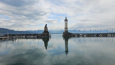 The image depicts the harbor entrance of Lindau (Bodensee), Germany, featuring the iconic Bavarian Lion statue on the left and the Lindau Lighthouse on the right. The harbor is filled with sailboats docked along a stone pier. Mountains are visible in the background, partially obscured by clouds. The water is calm, reflecting the sky and structures. The scene is captured during daylight hours.