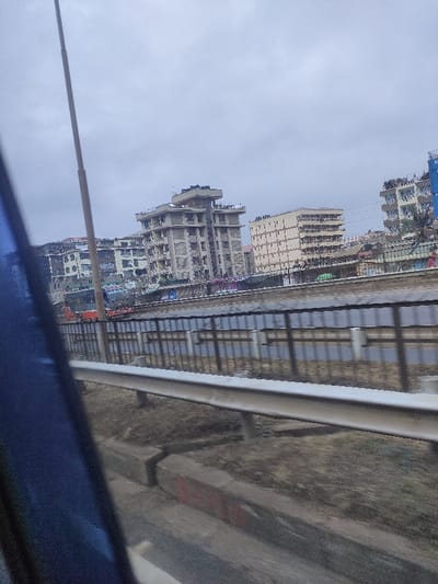 Buildings rise in the distance, several stories tall, under a cloudy sky. A guardrail and roadside infrastructure are in the foreground, suggesting a highway or elevated road view. The scene is slightly blurred, possibly taken from a moving vehicle, with the vehicle's window frame visible on the left.