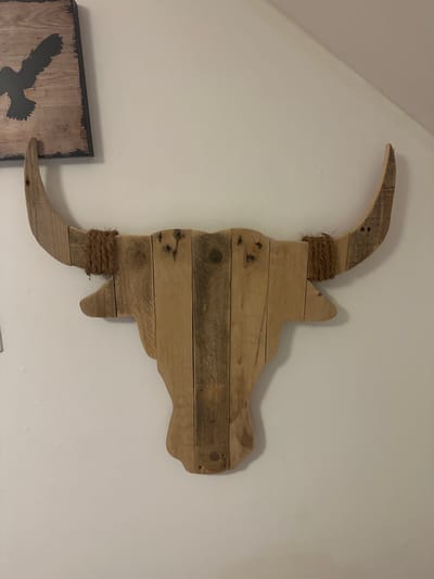 A wooden bull's head decoration is mounted on a white wall. The bull's head is made from several planks of wood, assembled to form the shape of a bull's head with prominent horns. The horns are wrapped with coarse brown twine near where they attach to the head. To the left of the bull's head, a framed wooden sign with a silhouette of a bird in flight is partially visible. The wall is painted a light color and appears to have some subtle texture. The overall scene suggests indoor home decor. This image was captured in Oldemarkt, Netherlands.