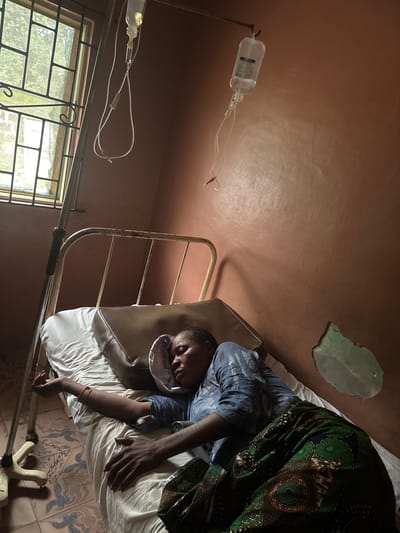 In a room in Ganyingbo, Nigeria, a woman lies on a hospital bed, asleep or unconscious. An IV drip bag hangs above her, its tube connected to her arm. She is wearing a blue patterned shirt and a green and brown patterned skirt. The room has a window with bars and a peeling, stained wall. The floor is tiled with a blue and white pattern.