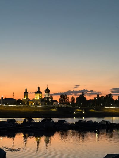 The image depicts a sunset view of the Spassky Monastery on the banks of the Volga River in Syzran, Russia. In the foreground, several boats are docked on the water, their dark silhouettes stark against the shimmering orange and blue reflections of the sky and the monastery. The monastery, illuminated by artificial lights, stands prominently on the horizon, its domes and towers silhouetted against the twilight sky. The sky transitions from a deep blue at the top to a warm orange hue near the horizon, with a few wispy clouds scattered across. The water's surface is rippled, creating a distorted reflection of the scene. The overall atmosphere is tranquil and picturesque, capturing the beauty of the city at dusk.