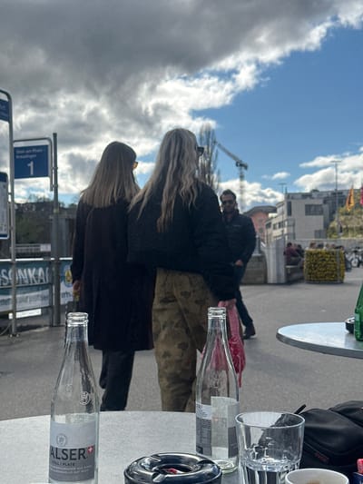 Here is a description of the image:

The image shows a street scene in Schaffhausen, Switzerland. A group of people is visible, including two women with blonde hair, and other blurred people. There is outdoor seating at a cafe or restaurant, with tables, glasses, and a water bottle labeled "Valser" and a metal ashtray visible. A sign indicating "Stein am Rhein - Kreuzlingen, 1" suggests the direction of travel. The sky is partly cloudy. There is a building with a yellow planter box filled with yellow flowers.