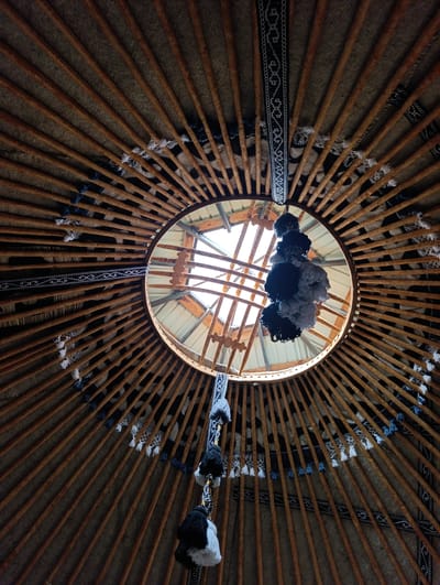 The image shows the interior ceiling of a yurt, a traditional nomadic dwelling. The yurt's roof structure is made of wooden poles radiating from a central circular opening, which appears to be a skylight. Decorative woven bands with geometric patterns in black and white are attached along the poles. Tassels in black, white, and blue hang from some of these bands, and also from a string that extends down from the central opening. A cluster of fluffy, dark and light-colored pom-poms hangs from a string directly beneath the skylight. The walls of the yurt, visible between the roof poles, are covered with a coarse, textured material, possibly felt or canvas. The image was taken from a low angle, looking upwards. The location is identified as Kegeti, Kyrgyzstan.