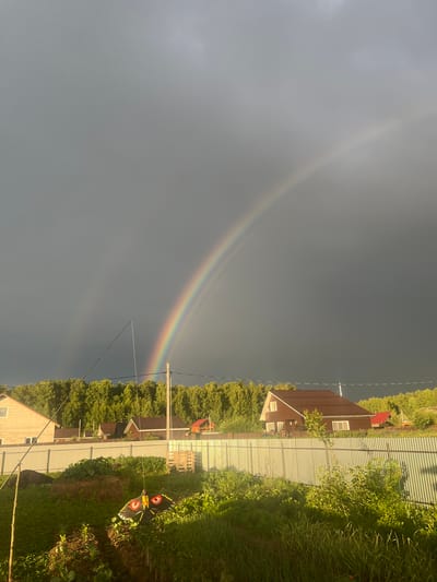 This media file captures a rural scene in Klepiki, Russia, likely during the late afternoon or early evening, as indicated by the soft, diffused lighting. The primary subjects are two houses, a corrugated metal fence, and lush greenery, suggesting a suburban or dacha setting. A prominent feature of the sky is a rainbow, arching across a gray, overcast sky, hinting at recent or ongoing rainfall. The rainbow's colors are vibrant, contrasting with the muted tones of the clouds and landscape.
To the right of the fence, a grassy area is visible, with some dirt patches and low-lying plants. Further back, trees form a dense backdrop, obscuring the distant horizon. The houses appear to be of typical rural construction, with pitched roofs and visible windows. One house has a dark brown exterior, while another closer to the foreground has a light-colored facade. The corrugated fence runs horizontally across the frame, separating the properties and creating a sense of enclosure.
A notable object in the foreground is a kite tethered to a pole, its colorful design adding a playful element to the scene. The kite has a black and yellow pattern with red eye-like markings. Electrical wires and poles are also visible, crisscrossing the sky and connecting the houses to utility infrastructure. The overall atmosphere is serene, with the rainbow suggesting a moment of natural beauty after a period of rain. No people are visible, and there is no discernible text in the image.