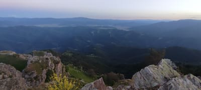The image shows a panoramic view of a mountainous landscape at dusk. In the foreground, there are rocky outcrops with sparse vegetation, including some yellow flowers. The middle ground and background are dominated by rolling hills and mountains covered in dark green forests. The sky is a pale gradient of blue and pink, indicating the transition from day to night. There are no people or man-made structures visible in the image, besides what appears to be a faint road or path on one of the rocky outcrops in the foreground. The location is identified as being in the city of Simeonovets, Bulgaria. The overall impression is one of natural beauty and serenity.