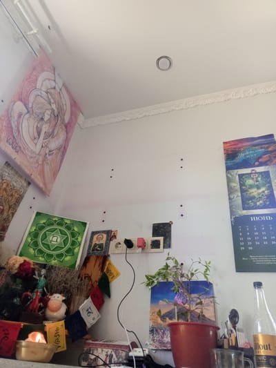 The image depicts an interior scene, likely a room, with a white wall as the dominant background. Several items are arranged on the wall and on a surface below it, suggesting a decorative or devotional space.
On the upper left wall, a large, pink and beige painting of two figures, possibly angels or spiritual beings, is displayed. Below this, on the wall, are what appear to be small holes, forming a vertical line. To the right of this, a calendar page is pinned to the wall, displaying the month of June with dates and some text in Russian. Above the calendar, a picture with a landscape and text in Russian is visible.
On a shelf or surface below the wall, various objects are arranged. A green square poster with a circular design is prominent. Next to it are several small framed images, one of which appears to be an icon. An electrical outlet with plugs and cables is also visible. To the right, a potted plant with green leaves is placed in front of another framed image of a mountainous landscape. Further to the right, a clear glass bottle with a yellow label is standing, and a metal cup is also present. Several small colorful flags with symbols are strung across this area. A lit candle in a brass bowl is in the foreground, on the left side of the shelf. Various other decorative items, including small figurines and dried plants, are also visible on the shelf.
The overall impression is that of a personal space decorated with spiritual or artistic items. The presence of an electrical outlet and a plant suggests it is a living space. The lighting appears to be natural, coming from an unseen source. The room is sparsely furnished, with the focus being on the wall decorations and the arrangement of items on the shelf. The location is not definitively identifiable as Пожарское сельское поселение, Ukraine, based on the visual information alone.