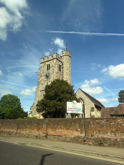 The image shows St. John's Stoke Church in Guildford, United Kingdom, viewed from a low angle beside a road. The church's stone tower rises into a blue sky with wispy clouds and contrails. A brick wall runs along the side of the road, in front of the church grounds. A sign for St. John's Stoke Church is visible, partially obscured by a tree. The road has double yellow lines.