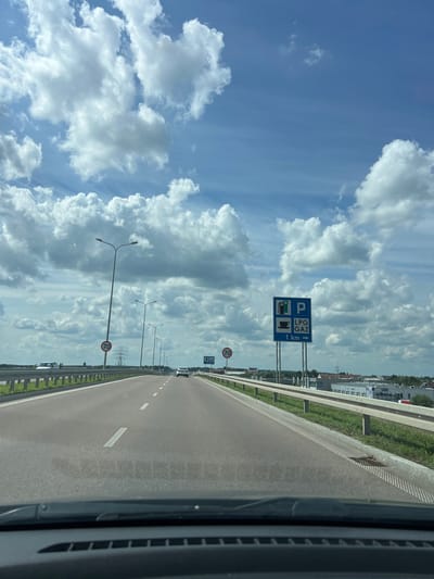 The image depicts a view from a vehicle traveling on a highway in Porosły-Kolonia, Poland. The road is paved with two lanes in the direction of travel, marked with white lines. A metal guardrail runs along the right side of the road. In the distance, several cars are visible on the highway. On the right side of the road, a blue road sign indicates "LPG GAZ" and a parking symbol with an arrow pointing to the right and the distance "1 km". There are also signs for a speed limit of 70 km/h. Tall streetlights are positioned along the road. The sky is filled with large, white cumulus clouds against a bright blue background. The landscape to the right of the road includes some industrial buildings and greenery. The image appears to have been taken during daylight hours.
