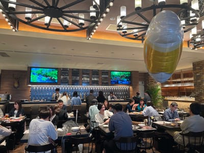 The image shows a restaurant interior with a large, oval-shaped, white and gold balloon suspended from the ceiling. The restaurant appears to be a gastropub with a long bar, rows of beer taps, and a large screen displaying a nature scene. The restaurant is filled with patrons and staff, and the tables are adorned with drinks, food, and plates. There is a mounted deer head on the wall above the bar, adding to the rustic atmosphere. The image is taken in a brightly lit environment with warm-toned lighting. The scene is bustling with activity, and patrons are engaged in conversation and enjoying their meals. The restaurant is located in Sumida City, Japan, at coordinates 35.71 latitude and 139.812 longitude.