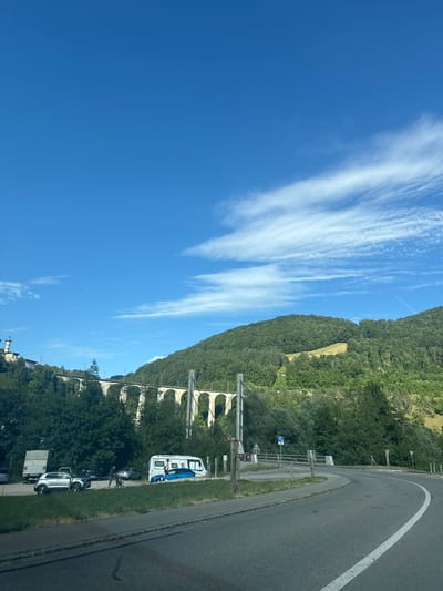 The image depicts a scenic view in Clos du Doubs, Switzerland, on a clear day with a vibrant blue sky and scattered wispy clouds. A large, arched viaduct, characteristic of railway infrastructure, spans across a lush, tree-covered hillside in the mid-ground. Below the viaduct, several vehicles are parked on a grassy area adjacent to a paved road. Among these vehicles are a white camper van with two individuals standing near it, a silver SUV, a dark-colored car, and a white truck. The road curves to the right in the foreground, leading away from the viewer. The environment is predominantly natural, with dense green foliage covering the rolling hills and mountains that dominate the background.