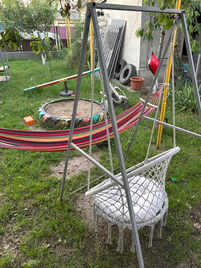 This is a medium-shot, outdoor, vertical photograph of a backyard scene in Pîrlița, Moldova. The foreground features a metal hammock stand with a striped hammock and a white net chair with a cushion. The hammock is multicolored with red, yellow, green, and purple stripes, while the net chair appears to be made of white rope and has a white cushion. The stand is constructed from gray metal poles and supports yellow and white ropes.
In the background, a circular tire filled with sand is visible, along with a red punching bag, some tires, and a terracotta pot. There are also some sheets of corrugated metal leaning against a wall. Green grass covers the ground, and some trees and foliage are present, suggesting a natural, lived-in outdoor space. The scene appears to be set during the daytime, with overcast weather, as indicated by the diffused light. The overall impression is one of a casual, possibly slightly unkempt, yard with recreational items. No people are visible in the frame.
There is some text visible on the red punching bag, but it is difficult to read clearly. The setting suggests a residential backyard, likely in a rural or suburban area of Moldova, given the location context.
