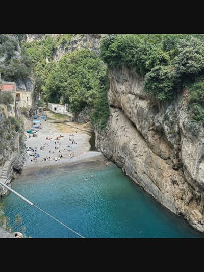 The image depicts a picturesque scene in the Fiordo di Furore, located near the town of Norcia, Italy, although the primary location is within the Amalfi Coast region, not Norcia itself. The scene is a narrow fjord with steep, rocky cliffs covered in lush green vegetation on either side. A small, pebble beach is visible where the fjord meets a body of clear, turquoise water.

Numerous people are scattered across the beach, some lounging on towels and chairs, while others appear to be walking or interacting. Several small boats are pulled up onto the shore. A set of stone stairs is carved into the cliff face on the left side of the image, providing access to the beach.

In the water, a few individuals are swimming and enjoying the cool, blue expanse. The overall impression is one of a secluded and tranquil natural oasis, popular for recreational activities. The architecture of a few buildings can be seen nestled into the cliffs on the left, adding a touch of human presence to the rugged landscape.