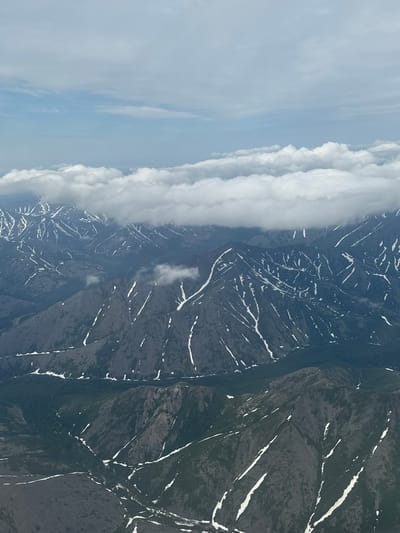 This aerial photograph captures a vast, rugged mountain landscape under a cloudy sky. The mountains are dark gray with patches of snow clinging to their slopes and ridges, giving them a streaked appearance. In the foreground and mid-ground, dense green forests cover the lower parts of some valleys and slopes. The sky above is a mix of pale blue and white clouds, with a prominent band of thick, white clouds obscuring the peaks of the furthest mountains. There are no people or man-made structures visible in the image. The scene depicts a remote, natural environment.