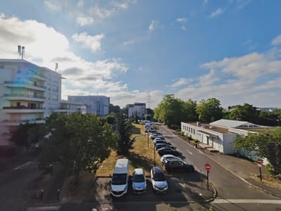 The image shows a street view in Gagny, France, on a clear day with some clouds. In the foreground, three vehicles are parked: a white van on the left, a silver hatchback in the middle, and a dark grey hatchback on the right. The street continues to the right, lined with several parked cars, including a black SUV and a silver sedan. A white building with a green roof and large windows is visible on the right side of the street, with a red and white circular "no entry" sign in front of it.
Further down the street, more parked cars are visible, as well as a white van and other vehicles further in the distance. Trees line the street, with a larger building visible in the background. The sky is blue with scattered white clouds, and the sun is visible, casting shadows. The overall scene depicts a residential or commercial area with parked vehicles and buildings.