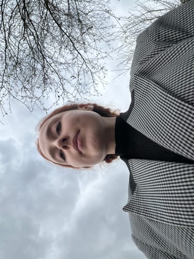 A low-angle photograph captures a young adult female from a perspective looking upwards. The subject has fair skin and reddish-brown hair, and is wearing a black ribbed high-neck top beneath a black-and-white checkered jacket. Her face is centered in the lower portion of the frame, with her gaze directed towards the camera. The background consists of a uniformly grey, overcast sky, against which the bare, dark branches of deciduous trees are visible across the upper section of the image. No other individuals or distinguishing landmarks are present in the frame. The scene is located in Eschau, France.
