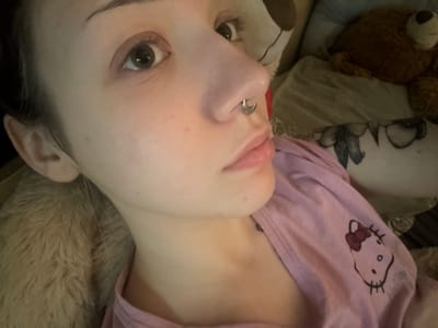 A close-up, eye-level, indoor shot shows the face of a pale-skinned person with dark hair, wearing a septum piercing. The person is looking slightly away from the camera, with their mouth slightly open. Their left eye is open and focused, while the right eye is partially visible. The person is wearing a pink t-shirt with a Hello Kitty graphic on it. The background is out of focus, but appears to be a bed with stuffed animals. The image is likely taken in a residential setting. There are no specific landmarks or environmental details to indicate the location in Olaine, Latvia.