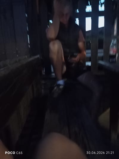 The image depicts a dimly lit, outdoor scene, likely in Kozichino, Bulgaria, at night. The central subject is a person, appearing to be male, sitting down with their hand on their head, suggesting a relaxed or contemplative pose. They are wearing dark clothing and what appear to be light-colored shoes with dark accents. The setting seems to be a rustic wooden structure, possibly a deck or a makeshift seating area, with dark wooden planks visible throughout the frame.
In the background, through gaps in the wooden structure, glimpses of illuminated rectangular shapes can be seen, possibly windows or lights from a building. A red bottle, likely a beverage, rests on a surface to the right of the central figure. The overall ambiance is dark, suggesting nighttime, and there are no clear indicators of weather conditions.
The image also contains visible text at the bottom left and right corners. The bottom left displays "POCO C65", indicating the model of the phone used to capture the image. The bottom right shows a date and time stamp: "30.06.2026 21:21".