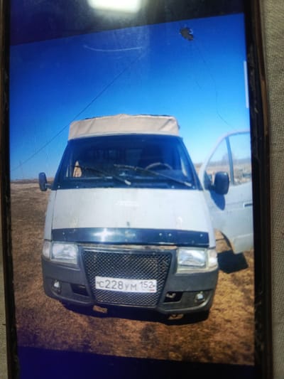 A white van with a beige roof is parked on a dry, grassy field under a clear blue sky. The van's driver's side door is open. A license plate on the front of the van reads "C228YM 152 RUS". The scene appears to be in an open, rural area, possibly on the outskirts of a city. There are no people visible in the image.