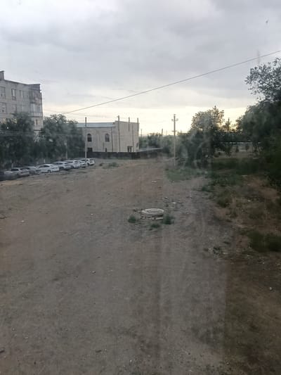 The image depicts a view of a city street in Satbayev, Kazakhstan. The scene is characterized by a dirt road in the foreground, with several parked cars lining the left side. In the background, there are buildings, trees, and utility poles. The sky is overcast with clouds.
The foreground is dominated by a dirt road with a manhole cover visible on the left side. The road is uneven and appears to be unpaved. To the left of the road, a row of parked cars is visible. These appear to be sedans and SUVs, mostly white and silver in color. Behind the cars, there are trees and a portion of a building.
The middle ground features more trees and a light-colored building with arched windows. This building appears to be a public or institutional structure. Utility poles and wires are also visible in this area, suggesting the presence of infrastructure.
The background is primarily filled with trees and the sky. The sky is cloudy, with a hint of light breaking through the clouds in the distance, suggesting either dawn or dusk. The overall impression is a somewhat utilitarian urban environment.