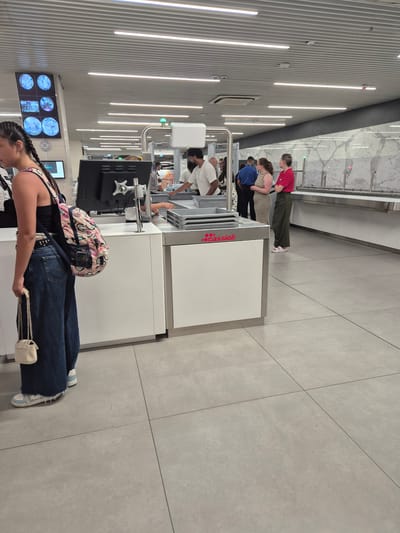 The image depicts an interior scene at Luqa Airport, Malta, likely within the security screening area. In the foreground, a young woman with braided hair is facing away from the camera, wearing a black tank top, dark blue wide-leg jeans, and white sneakers. She is carrying a backpack with a pink and white cartoon pattern and a small white handbag with a chain strap. To her left, a monitor displays multiple circular images, possibly from security cameras.
In the mid-ground, a security checkpoint is visible. A man in a white striped shirt is standing behind a counter, interacting with an individual whose hands are on a conveyor belt. To the right of the man, two women are standing in a line; one is wearing a striped dress, and the other is wearing a red top and green pants. Behind them, a long counter with a marble-patterned backsplash extends into the background. The floor is tiled with light gray square tiles. Overhead, white linear lights illuminate the space, and a metallic ceiling with horizontal slats is visible. The overall environment appears clean and functional, consistent with an airport's security processing area.