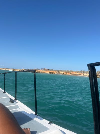 The image shows a view from a boat, likely a small watercraft, in Unknown, Venezuela. A clear blue sky dominates the left half of the frame. A body of water, possibly the ocean or a large lake, fills the right half, with land visible in the distance. The coastline features a combination of sandy areas and sparse vegetation, mainly cacti. A portion of the boat's structure, including railing and deck, is visible in the foreground. A human leg and foot are also visible.