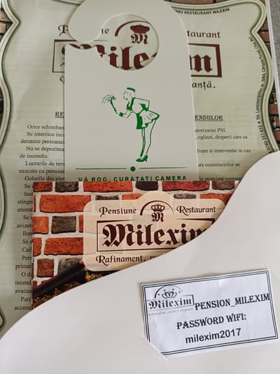 This media file captures a close-up view of a hotel room door hanger and a sheet of paper displaying Wi-Fi information. The door hanger, made of white cardstock, features a stylized illustration of a person dressed in green, holding a bouquet of flowers. It also includes text related to hotel policies and restaurant information. A logo with the name "Milexim" is prominently displayed.
The Wi-Fi information sheet is a small, rectangular piece of paper with a decorative border. It shows the hotel's name, "PENSION MILEXIM," and the Wi-Fi password, "milexim2017." The "Milexim" logo is repeated on this sheet.
In the background, a textured surface resembling a brick wall is visible, likely part of the hotel's interior decor. The overall impression is that of a hospitality establishment, possibly a hotel or guesthouse, aiming to provide guests with essential information and a welcoming atmosphere. The lighting appears to be indoor and even, suggesting the photo was taken inside. The text is primarily in Romanian, indicating the location is likely in Romania. The address visible on a faded label mentions "Str. Laminoriştilor, nr. 208A, Câmpia Turzii, Cluj, RO," confirming the location context.