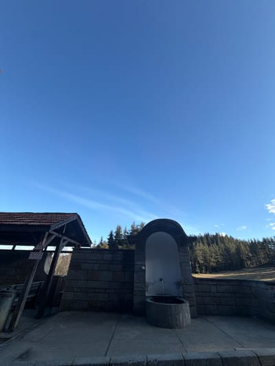 The image depicts an outdoor scene in Kasak, Bulgaria, under a clear blue sky with faint, wispy white clouds. In the foreground, a paved area leads to two main structures. To the left, a wooden shelter with a dark brown tiled roof is visible, featuring wooden support beams and an obscured sign with Bulgarian text. To the right of the shelter, a stone wall constructed of grey rectangular blocks supports a fountain. The fountain consists of an arched alcove with a light-colored interior, from which a metal spout emits a steady stream of water into a semi-circular stone basin below. No individuals are present. In the background, a dense evergreen forest extends across the horizon, bordered by a dry, grassy slope immediately behind the stone wall.