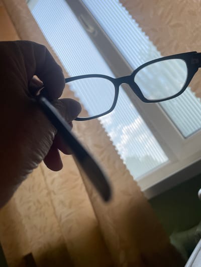 A hand holds black-rimmed glasses in front of a window with vertical blinds. The background shows a window with white blinds and a glimpse of what appears to be a curtain. The scene is indoors, likely in a residential setting. The image does not provide any specific details that would allow for the determination of its location within the city of Tikhvin, Russia.