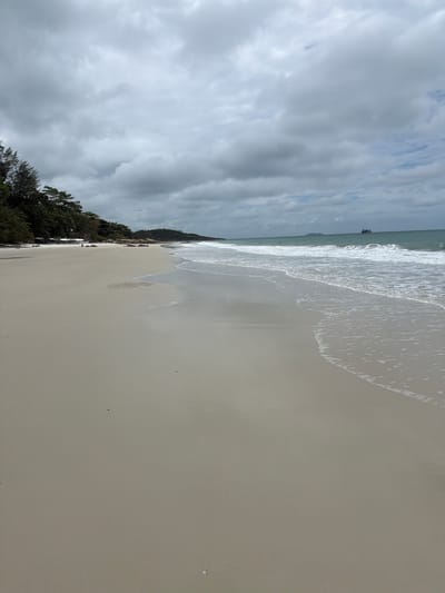 The image depicts a serene beach scene in Ao Hin Khok, Thailand, under a cloudy sky. The foreground is dominated by a wide expanse of pale, smooth sand, wet near the water's edge where gentle waves are breaking. The sea is a muted turquoise, with white foam marking the surf. In the distance, a small boat can be seen on the horizon. Along the left side of the frame, a line of lush green trees separates the beach from what appears to be a small settlement with white structures, possibly umbrellas or temporary shelters. A few scattered rocks are visible on the sand further down the beach. The overall atmosphere is calm and slightly overcast, suggesting a peaceful day at the coast.