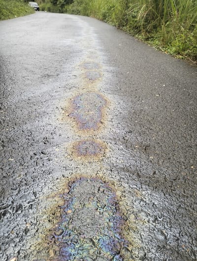 The image shows a close-up of an asphalt road with a rainbow-colored oil slick. The road is wet, and the oil slick reflects light in iridescent patterns of purple, blue, green, yellow, and orange. The road surface is textured with small stones and appears to be slightly uneven. In the background, the road curves to the left, and on the right side of the road, there is tall, green grass and vegetation. In the distance on the left, a dark-colored vehicle is visible on the road. The scene is outdoors, and the lighting suggests it is daytime. The image does not contain any people or obvious signs of recent activity beyond the presence of the oil slick and the vehicle. The location is identified as Aluepapeun, Indonesia.