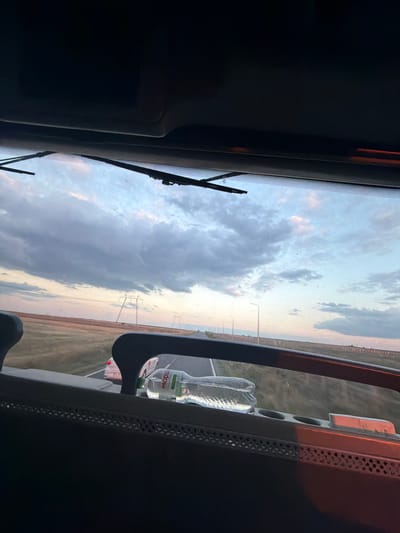 The image is taken from inside a vehicle, looking out through the windshield. The sky is filled with clouds in shades of gray and pink, suggesting either sunrise or sunset. The landscape outside appears to be a flat, open area with fields of dry grass or crops and a road stretching into the distance. Power lines are visible on the horizon.
In the foreground, inside the vehicle, a plastic water bottle is visible, resting on a dashboard or console. The bottle has a green label with white text that is partially obscured. Part of the vehicle's interior, including a dark-colored frame or dashboard element, is also visible. A portion of another vehicle, likely a car, can be seen on the road outside.
The scene suggests a journey through a rural or semi-rural area, likely on a highway or main road, during twilight. The location is specified as Unknown, Romania.