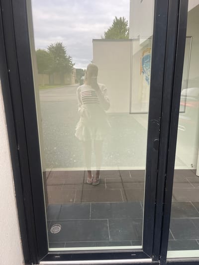 A reflection in a glass window shows a person wearing a white outfit with an American flag design. The reflection also shows trees, buildings, and a cloudy sky. The window frame is black. The reflection overlays a view of tiled flooring and interior elements of a building, including framed artwork. The photograph is taken at an angle.