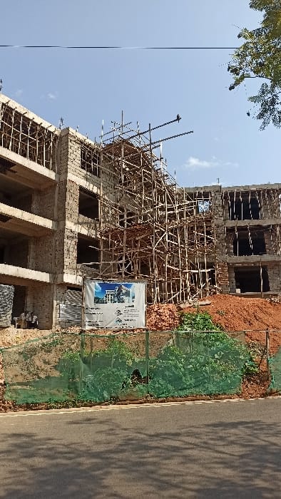The scene depicts a multi-story building under construction in Kigali City, Rwanda, viewed from an asphalt road. The structure features a concrete frame with partially completed cinder block walls. Extensive wooden scaffolding covers a significant portion of the central and right sections of the building. Several individuals, likely construction workers, are visible on the ground level of the building's left side, appearing to be resting or conversing.
In front of the building, a large white banner displays a rendering of the finished structure and project information. To the right, a pile of red bricks is present on the reddish-brown soil of the sloped terrain. A green mesh fence runs along the immediate foreground, separating the construction site from the road. The asphalt road itself shows tree shadows. The sky is clear blue with minimal clouds, indicating a bright, sunny day. A bird is perched on a scaffolding pole in the upper right quadrant of the building. Tree foliage is visible in the top right corner of the image.