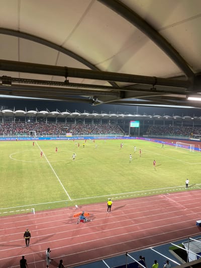 A stadium hosts a soccer match, viewed from an elevated position beneath a structural canopy. The field is green with visible white lines and players in red and white uniforms. A running track surrounds the field. Spectators fill the stands. A stretcher with medical personnel is visible on the track.