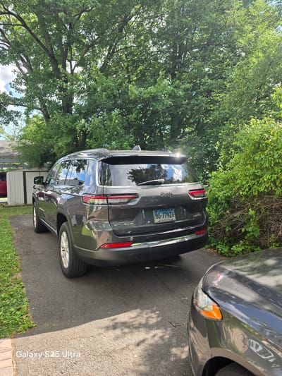 In West Hartford, Connecticut, USA, a dark grey Jeep Grand Cherokee is parked on a paved driveway. The vehicle, bearing the license plate "BU-77926," is positioned at an angle, with its rear end closer to the camera. The front section of another dark grey vehicle is visible in the foreground on the right.
The scene is set in a residential area with a lush green tree providing shade and a dense green bush bordering the driveway on the right. A white shed with a sloped roof and a smaller red vehicle can be seen in the background on the left. The sky is partly cloudy, with clear blue patches visible through the tree branches. The overall environment suggests a sunny day in a suburban setting.