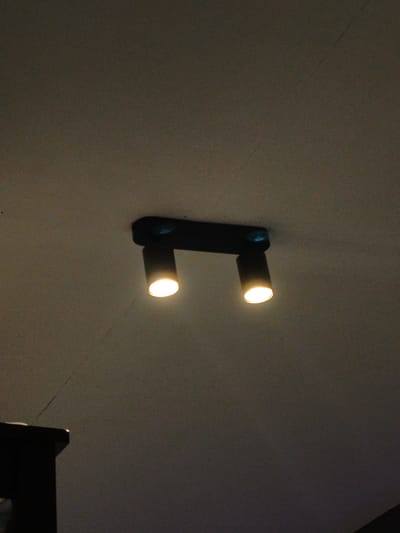 A ceiling-mounted black track light fixture with two cylindrical spotlights is visible against a dark, textured ceiling. Both spotlights are illuminated, casting a warm yellow light. The fixture is positioned centrally in the frame, with a portion of a dark, furniture-like object visible in the lower left corner of the image. The scene appears to be indoors, in a room with low ambient light. The image quality is grainy, suggesting it was taken in low-light conditions. The location of Cavite City, Philippines is not discernible from the visual information provided in the image.