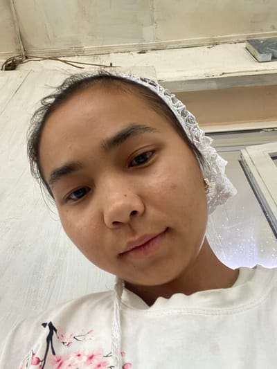 A close-up, low-angle, waist-up shot captures a young person with dark hair and brown eyes, wearing a white top with a floral print and a white lace headband. The subject's skin appears to have minor blemishes. Behind the subject, a white wall with textured paint is visible. To the right, a window with white framing and some reflection can be seen. The overall lighting suggests an indoor setting. The environment does not provide any specific clues to pinpoint a location within the city of Kyzyl-Kyya, Kyrgyzstan.