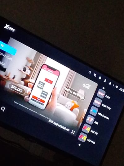 The image shows a television screen displaying a streaming service interface. On the left side of the screen, there are menu options labeled "TV" and "DESTACADOS". Below these options, there is a button with the text "SALDO" highlighted in white. In the center of the screen, a hand is holding a smartphone displaying a payment confirmation screen. To the right of the smartphone, a graphic with the word "dish" is visible, followed by the text "Fecha de pago". Further to the right, a list of channels is displayed, including "ECOF", "ECOF FHD", "NBA Eventos", "A3S", "A&E HD", and "A&E FHD". At the bottom right corner of the screen, the time "23:53" is visible. The background of the television screen shows a blurred interior scene with furniture. The television itself is dark and reflects some light from the room. The overall scene is a close-up of a television screen, likely in a home environment in Atamo Sur, Venezuela, displaying a user interface for a television or streaming service.