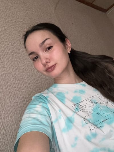 A woman lies on a beige carpet, head tilted towards the camera. She has long, dark hair, light skin, and wears small earrings. She's dressed in a white t-shirt with light blue tie-dye patterns and a graphic design featuring Japanese text. The left side of her body faces the camera. A portion of a doorframe is visible in the background.