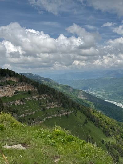 The image shows a vast mountain landscape under a cloudy sky. In the foreground, there is a steep, grassy slope dotted with small white and yellow wildflowers. A large rock is visible on the left side of the foreground. The middle ground is dominated by a rocky cliff face with layers of exposed stone, covered in patches of green grass and dotted with evergreen trees. Below the cliff, the slope continues downwards, covered in dense green vegetation and trees. In the background, a wide valley stretches out, with a river or body of water visible in the distance. Further back, multiple layers of hazy green mountains recede into the distance, with the sky above filled with large, white, puffy clouds against a pale blue background. The scene depicts a natural, rugged mountainous environment, characteristic of the region near Nizhnyaya Teberda, Russia, a town located in the Karachay-Cherkess Republic, known for its mountainous terrain and natural beauty. There are no people or man-made structures immediately visible in the main focus of the image, suggesting a remote, natural setting.
