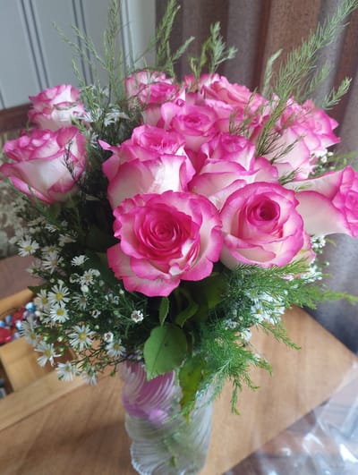 A close-up, eye-level shot captures a bouquet of pink and white roses, interspersed with delicate white filler flowers and feathery green foliage. The roses, with their vibrant pink edges fading to white centers, are the primary focus. The bouquet is arranged in a clear, fluted glass vase. The vase sits on a wooden surface, likely a table, with a blurred background that suggests an indoor setting. To the left, a glimpse of a wooden tray containing colorful, wrapped candies is visible. The overall impression is one of a carefully arranged floral gift, possibly for a special occasion. There is no indication of the city of Hrazdan, Armenia, or any other specific location within the image.