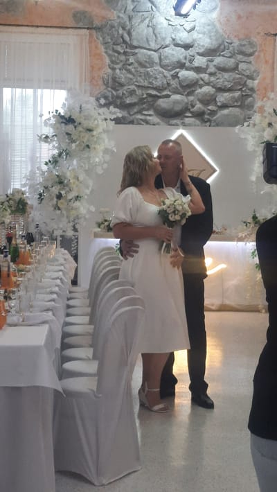 A wedding ceremony is taking place indoors in a room with a stone wall in the background. A bride, wearing a white dress and holding a bouquet, is embracing and kissing the groom, who is in a dark suit. The room is decorated with white floral arrangements and a neon sign in the shape of a diamond. A long table set for a meal with white tablecloths, chairs covered in white fabric, and bottles of drinks is visible on the left side of the image. The floor is a light-colored speckled tile. The overall atmosphere is celebratory. This event appears to be occurring in or near the village of Zhodzishkava, in the Zhodzishkava rural council area of Belarus.