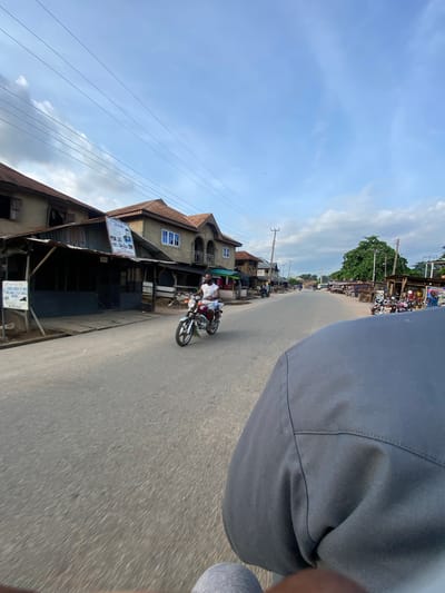 This image depicts a street scene in Igarra, Nigeria, captured from the perspective of a passenger in a moving vehicle, with the blurred road surface visible in the lower frame and the top of a grey-clad leg and shoulder in the immediate foreground. An asphalt road, slightly wet in patches, extends into the distance.

On the left side of the road, a series of multi-story buildings with light-colored walls and reddish-brown corrugated metal roofs are visible. One prominent building features an attached open-sided structure with a corrugated metal canopy, above which hangs a large white and blue banner displaying text, including "FRIDAY & CO.", and several photographic portraits. Closer to the foreground on the left, a white sign with black text is partially visible. Utility poles and a dense network of overhead wires run parallel to the street.

In the middle ground, a man wearing a white sleeveless shirt and dark shorts is riding a silver motorcycle with red accents, traveling towards the camera on the opposing lane.

Further down the street on the right side, more buildings similar in style, some with blue-framed windows and balconies, line the road. A large, lush green tree stands prominently beyond these structures. Numerous motorcycles are parked along the right roadside, and several people are observed standing or sitting nearby. The sky above is a mix of clear blue and scattered white to light grey clouds, with more overhead wires visible. The overall scene suggests a moderately active commercial or residential street.