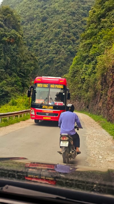 A medium shot photograph taken from a vehicle traveling on a narrow road in Cán Tỷ Commune, Vietnam, depicts a red bus heading towards the camera and a man on a motorcycle in front of the camera vehicle. The bus, labeled "MÈO VẠC HẢI PHÒNG" and "HÙNG LONG," has the license plate "23A-01253." The motorcyclist wears a blue shirt and a helmet; the motorcycle's license plate reads "23-L1 093.60." Lush green hills and cliffs border the road on both sides. The bus's reflection is visible on the vehicle's dashboard from which the image is being taken.