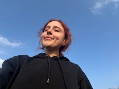 A low-angle, upward-facing shot captures an individual outdoors. The subject, a young person with reddish-brown hair styled loosely around their face, is positioned in the lower portion of the frame, facing slightly upward and directly towards the camera. They exhibit a subtle smile, with visible dimples on their cheeks. The individual wears a black hooded sweatshirt with drawstring cords featuring silver metallic tips. One earlobe shows a small, circular ear piercing. The background consists predominantly of a clear, bright blue sky with a few scattered, wispy white clouds. No specific landmarks or distinguishing ground features are visible to determine a precise location within the city of Toul, France. The scene suggests a daytime setting with ample natural light.