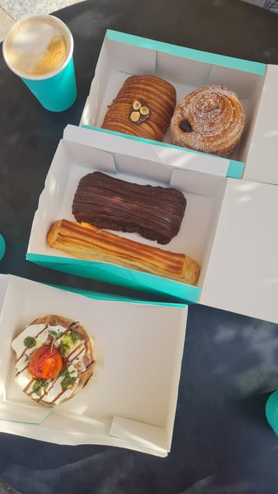 The image captures a selection of pastries and a coffee, neatly arranged in three white cardboard boxes with turquoise accents, set upon a dark table or surface. The scene appears to be well-lit by natural light, suggesting daytime in Kiryat Gat, Israel.

In the leftmost box, there are two sweet pastries. The upper one is round, covered in powdered sugar, and has a dark filling visible in its center, resembling a cinnamon roll or a pain aux raisins. Below it, a golden-brown, layered pastry, possibly a pain au chocolat, is topped with a chocolate drizzle and three whole hazelnuts.

The middle box contains two rectangular pastries. One is a rich, dark brown with prominent ridges, indicating a chocolate-infused pastry. Beside it is a lighter, golden-brown, elongated pastry, possibly a plain puff pastry stick or a variant of a croissant.

The rightmost box holds a single savory tart or pastry. It is circular, featuring a topping of melted white cheese, a roasted red cherry tomato, dollops of green pesto or herbs, and a dark, glossy balsamic glaze drizzle.

To the bottom left of the frame, a turquoise paper cup contains a frothy latte or cappuccino, with distinct layers of coffee and milk foam visible. The bright lighting creates soft shadows on the table, contributing to a clear and inviting presentation. No people are visible in the image, and no legible text can be identified on any of the items or packaging. The arrangement suggests a casual and pleasant meal or coffee break.